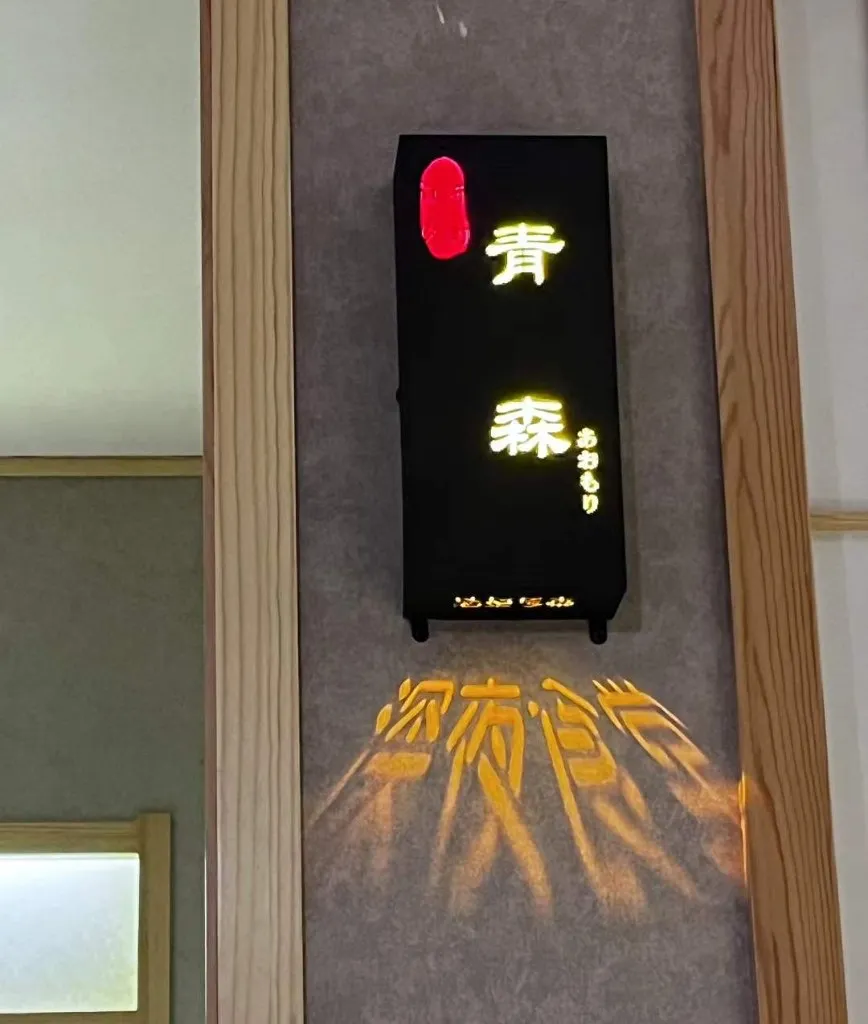 日式餐厅墙上的黑色灯箱招牌，暖黄的「青森 あおもり」字样亮着，左上角一枚红色灯饰，灯光在灰色墙面上投下一层重影