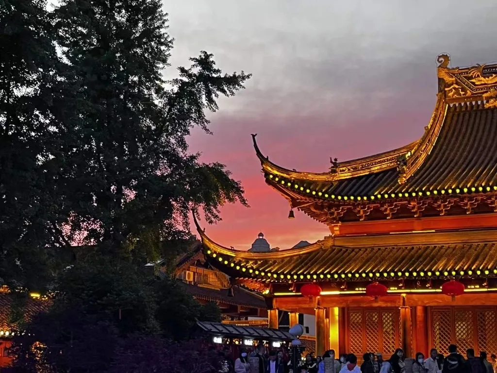 暮色里的中式寺庙，金色琉璃屋檐与翘起的檐角亮着灯带，屋下挂着红灯笼，天空是粉红与青灰交界的晚霞，殿前站满了人