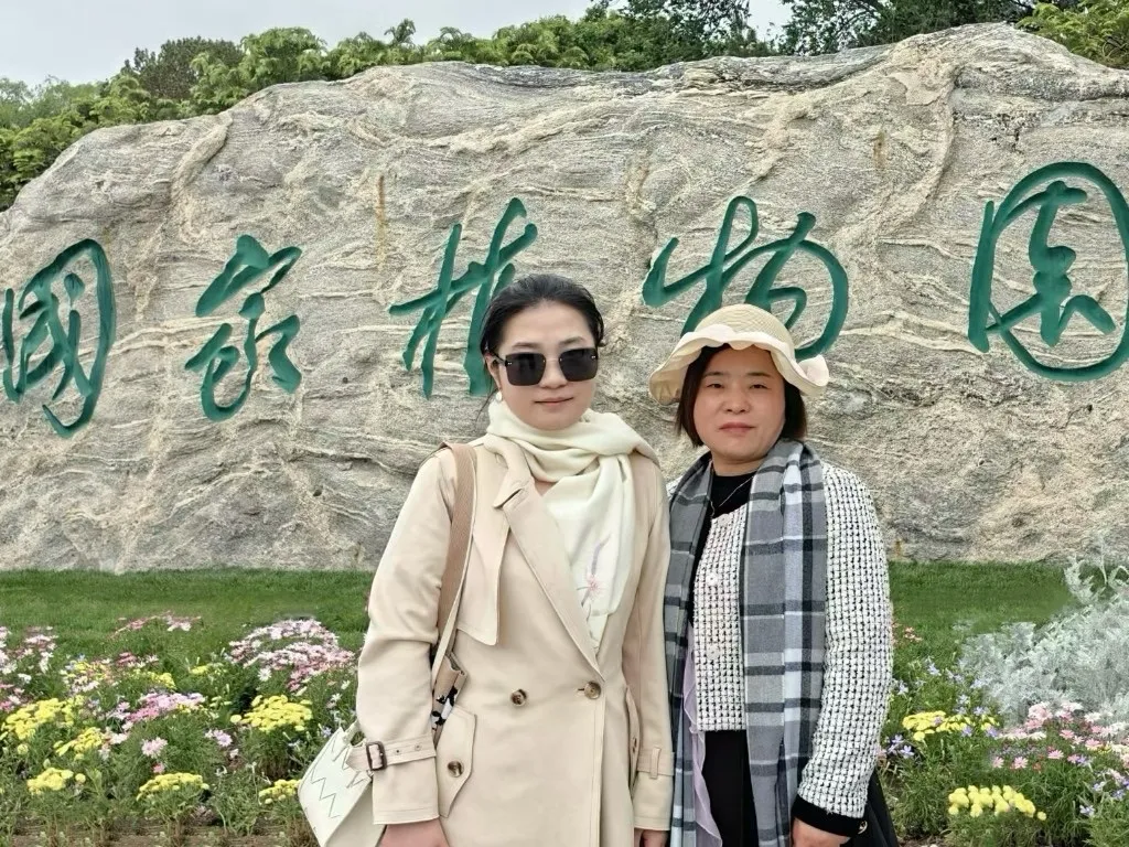 国家植物园的题名石前，两个人并肩站着合影，身前是一片黄白色的菊花